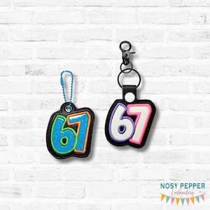 Può includere: Due portachiavi con il numero 67 ricamato. Uno ha un design blu e arancione, l'altro bianco e rosa. Entrambi i portachiavi hanno un bordo nero e un fermaglio in metallo.