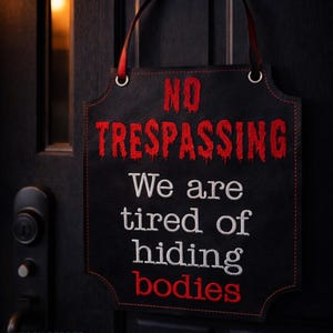 May include: A black sign with red and white embroidered text. The sign reads "NO TRESPASSING We are tired of hiding bodies." It has a red ribbon for hanging and is attached to a black door.