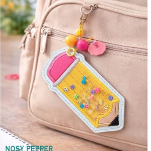 May include: A yellow pencil-shaped keychain with a pink eraser and a clear center filled with colorful confetti and smiley face beads. The keychain is attached to a beige backpack with a decorative beaded charm.