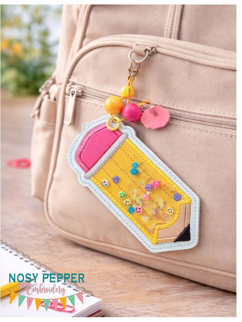 May include: A yellow pencil-shaped keyring with a pink eraser and a clear centre filled with colourful confetti and smiley face beads. The keyring is attached to a beige backpack with a decorative beaded charm.