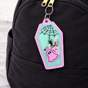 May include: A Halloween-themed keychain in the shape of a coffin, featuring a pink ghost, spider, and web design. The keychain is attached to a black backpack with a gold zipper.