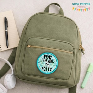 Peut inclure: Un sac à dos vert olive avec une poche avant zippée et un écusson rond. L'écusson porte le texte "PRAY FOR ME. I'M PETTY" en noir et bleu sarcelle. Un carnet, un stylo, un casque et un surligneur sont également visibles.