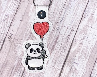 May include: A white leather keychain with a black and white panda holding a red heart-shaped balloon.