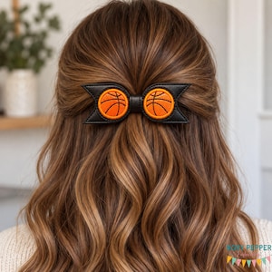 May include: A black hair bow featuring two circular orange basketball patches with black stitched lines. The bow has a structured, pointed design and is centered in the hair. The basketballs are embroidered with orange thread and black details.