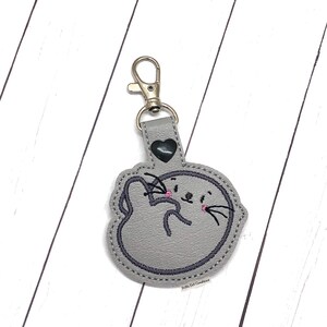 May include: Gray felt keychain with a cute cartoon seal design. The seal has black eyes, a pink nose, and a black mustache. The keychain has a silver metal clasp.