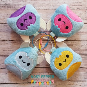 Puede incluir: Cuatro monstruos de peluche azul y blanco con caras de diferentes colores. Los monstruos tienen alas blancas y están dispuestos en círculo. Las caras son moradas, rosas, amarillas y azules.