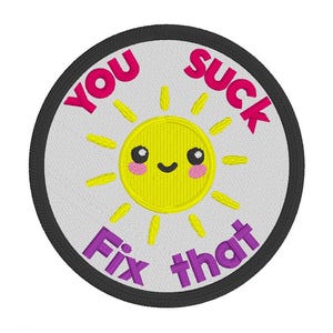 May include: Embroidered patch with a white background and a black border. The patch features a yellow smiling sun with pink cheeks and the phrases "YOU SUCK" and "Fix that" in pink and purple lettering.