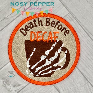 May include: A round, orange-bordered patch with a beige background. The patch features a brown coffee cup with a skeleton hand holding it. The text "Death Before Decaf" is written in a black cursive font above the cup.