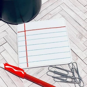 以下が含まれることがあります： ノートのページのような、赤と青の線が引かれた白い正方形のコースター。コースターは、黒いマグカップと赤いペンのある明るい木製の表面にあります。