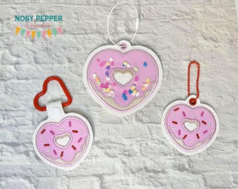 Può includere: Tre portachiavi e portabiglietti a forma di cuore. I disegni rosa presentano un tema a forma di ciambella con zuccherini e un centro a forma di cuore. Un portachiavi ha una chiusura rossa a forma di cuore, un altro un anello bianco e il terzo una catena rossa.