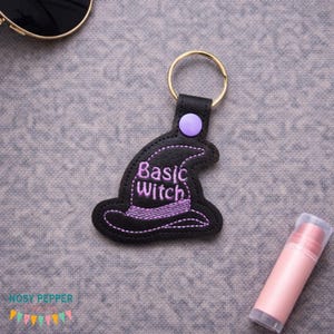 Peut inclure: Porte-clés en forme de chapeau de sorcière noir avec l'inscription "Basic Witch" brodée en violet. Le porte-clés a un anneau doré et un bouton violet. Un baume à lèvres rose est également visible.