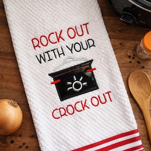 Peut inclure: Serviette de cuisine blanche avec texte brodé rouge et noir et un motif de mijoteuse. Le texte dit "ROCK OUT WITH YOUR CROCK OUT". La serviette a une bordure rayée rouge et blanche et un motif texturé.