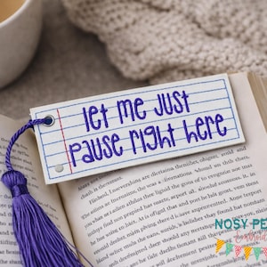 May include: A bookmark with the text "Let me just pause right here" in blue, resembling a lined notebook page. It has a purple tassel and is placed in an open book.