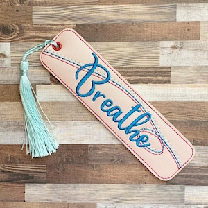 Peut inclure: Un marque-page en cuir rose avec le mot "Breathe" brodé en bleu et un pompon bleu clair.