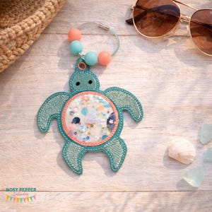 May include: A teal turtle-shaped keychain with a clear center filled with colorful confetti and small embellishments. The keychain has a beaded accent with pink and blue beads. Sunglasses and a woven bag are in the background.