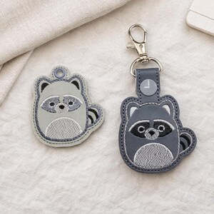 May include: Two raccoon-themed keychains. One is a flat, gray embroidered patch with a ring for attaching to a keychain. The other is a gray leather keychain with a silver clasp. Both have a raccoon design.