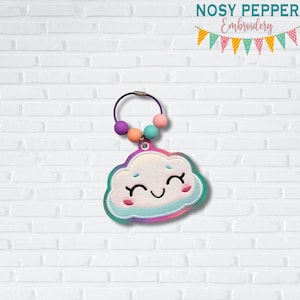 Op de afbeelding: Een wolk-vormige geborduurde sleutelhanger met een lachend gezicht, regenboogafwerking en een paarse lus met kleurrijke kralen. De sleutelhanger heeft de tekst "NOSY PEPPER Embroidery".