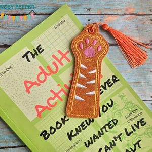 May include: Orange and white tiger paw bookmark with a pink paw pad and a tassel. The bookmark is on top of a book with the text "The Adult Achieved Book Knew You Wanted" on the cover.