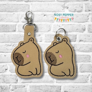 May include: Two brown capybara keyrings with embroidered details. The capybaras are sitting with their eyes closed and have pink noses. The keyrings are attached to silver key rings.