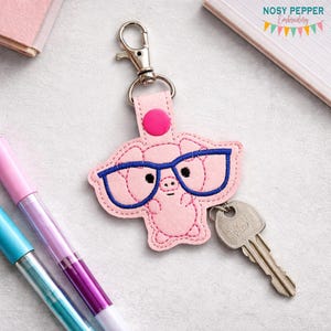 May include: A pink pig-shaped keychain with blue glasses and a silver key attached. The keychain has a pink button and a silver clasp. The pig has black eyes and is made of stitched fabric. The keychain is a fun accessory.