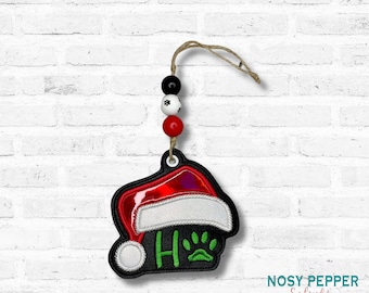 May include: A Christmas ornament featuring a Santa hat design with the word "HO" in green and a paw print. The ornament is black with red and white accents, and is strung with beads in red, black, and white. A jute string is attached for hanging.