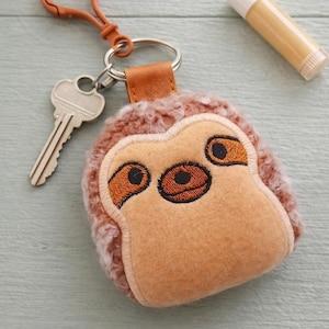 Puede incluir: Un llavero de perezoso de peluche con una correa de cuero marrón y una llave plateada. El perezoso tiene ojos y nariz bordados. Un tubo de bálsamo labial está junto al llavero. Un accesorio divertido.
