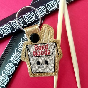 May include: A gold-coloured leather keychain shaped like a Chinese takeaway container with the text "Send Noodles" and a smiling face. The keychain is attached to a silver key ring and is next to two wooden chopsticks.