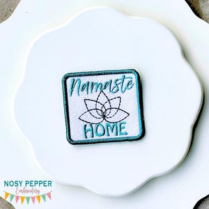 May include: Square embroidered patch with a teal border and the words "Namaste HOME" in teal script. A black outline of a lotus flower is centered between the words. The patch is on a white, scalloped surface.