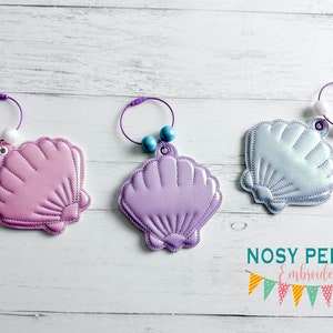 Może przedstawiać: Trzy różne kolorowe muszle wykonane z tkaniny z przymocowanymi fioletowymi kółkami i koralikami. Muszle są różowe, fioletowe i jasnoniebieskie. Tekst "NOSY PEPPER Embroidery" znajduje się na dole obrazu.