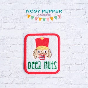 May include: Embroidered patch featuring a cartoon nutcracker soldier with a red hat and the text "Deez Nuts" in green. The patch has a red border and is set against a white background.