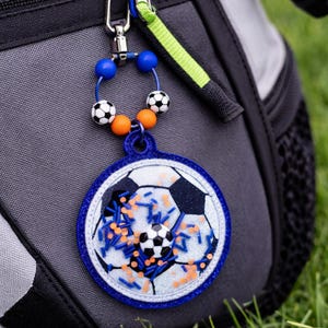 May include: A football-themed keyring with a blue and white border, featuring a football design with blue and orange accents. The keyring is attached to a backpack with a silver clasp and a beaded accent.