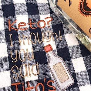 May include: A white and blue checkered kitchen towel with the text "Keto? I thought you said Tito's" embroidered in brown and orange thread. A bottle of Tito's Vodka is partially visible in the background.