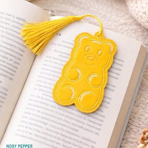 Könnte beinhalten: Ein gelber Gummibärchen-Lesezeichen mit weißen Akzenten und einer passenden Quaste. Das Lesezeichen liegt auf einem offenen Buch. Der Text "Nosy Pepper Embroidery" ist unten sichtbar.