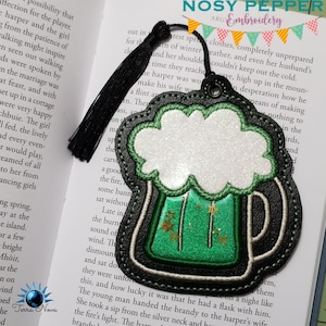 May include: A black leather bookmark with a green beer mug design. The mug has a white foam top and gold stars. The bookmark has a black tassel.