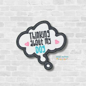 Op de afbeelding: Een witte, wolkvormige patch met zwarte contouren en blauwe en witte tekst die "Thinking about my dog" zegt, met twee roze harten aan weerszijden van de tekst. De patch bevindt zich op een witte bakstenen muur.