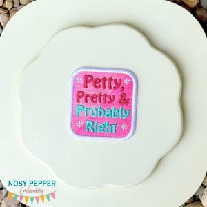 Pode incluir: Patch bordado rosa e turquesa com a frase «Petty, Pretty & Probably Right». O patch quadrado é decorado com flocos de neve e está sobre um fundo branco com vieiras. O logotipo «Nosy Pepper Embroidery» está na parte inferior.