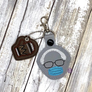 May include: A gray leather keychain with a white embroidered face wearing glasses and a blue face mask. The keychain has a brown leather heart-shaped charm with a white embroidered design.