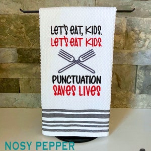 May include: White kitchen towel with gray stripes. The towel has embroidered text that reads "Let's eat, kids. Let's eat kids. Punctuation saves lives." with a pair of crossed forks.
