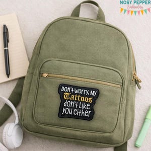 May include: Olive green backpack with a gold zipper and a patch that reads "Don't worry, my tattoos don't like you either." A black pen, notepad, and headphones are visible in the background. The backpack has a small front pocket.