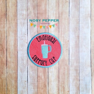 May include: A round, red embroidered patch with blue stitching. The patch features a white cup with a straw and the text "Emotional Support Cup".