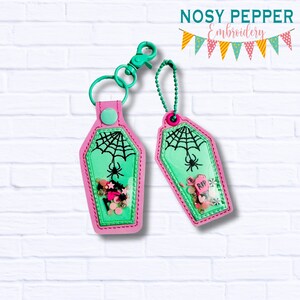 Puede incluir: Dos llaveros en forma de ataúd rosa y verde con un diseño de telaraña y araña. Los llaveros tienen ventanas de plástico transparente llenas de purpurina y abalorios coloridos. El texto "RIP" es visible en uno de los abalorios.