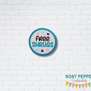May include: A white circle embroidered patch with a blue border and the text "free shrugs" in black. The patch is surrounded by small pink, blue, and purple hearts.