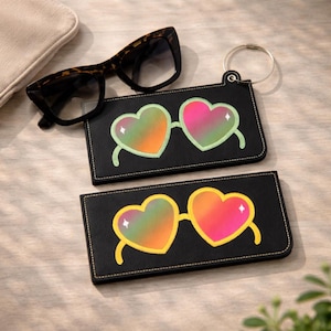 May include: Two black eyeglass cases with heart-shaped sunglasses designs. One case has a green outline, the other has a yellow outline. Tortoiseshell sunglasses rest on top. A silver ring is attached to one case.