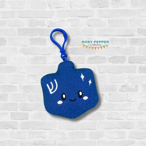 May include: A blue plush dreidel keychain with a white Hebrew letter and a smiling face. The dreidel has a blue plastic clip attached to it.