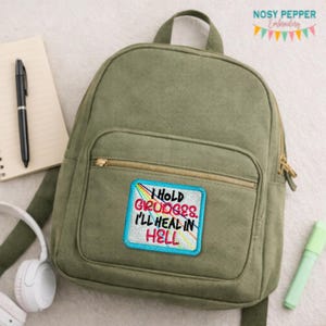 May include: An olive green backpack with a front zip pocket and a top handle. The backpack has a rectangular patch embroidered with the text "I HOLD GRUDGES I'LL HEAL IN HELL". A pen, notepad, headphones, and a highlighter are also visible.