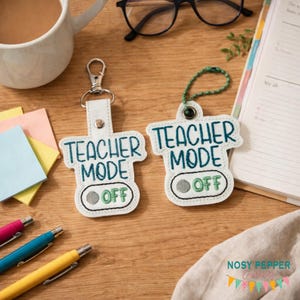 以下が含まれることがあります： 「TEACHER MODE OFF」の文字がターコイズブルーの刺繍で施された2つの白いキーホルダー。1つはシルバーの留め金、もう1つはグリーンのループが付いています。木製の表面にペン、マグカップ、プランナーと並んでいます。