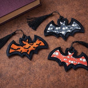 May include: Three bat-shaped bookmarks with black tassels. One is orange with black bat silhouettes, another is red with white eye patterns, and the third is gray with white eye patterns. A book is in the background.