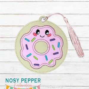 May include: A round, embroidered bookmark featuring a smiling pink donut with colorful sprinkles. The bookmark has a light pink tassel and a metal grommet for hanging. The text "NOSY PEPPER Embroidery" is at the bottom.