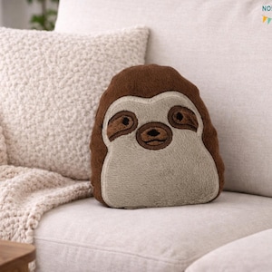 Puede incluir: Un cojín con forma de perezoso, en tonos marrón y beige, con detalles faciales bordados y una textura suave. Descansa sobre un sofá blanco, con una almohada y una manta texturizadas en el fondo. El cojín mide unos 25 cm de altura.