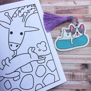 以下が含まれることがあります： キリンと、その背中に横たわる小さな動物の白黒のイラスト。紫色のタッセルと、青い浮き輪の上で横になっている白黒のフレンチブルドッグの付いたしおり。